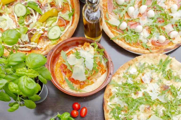 Overhead view of four pizzas with fresh basil, mozzarella, and assorted toppings arranged on a dark surface with herbs and vegetables