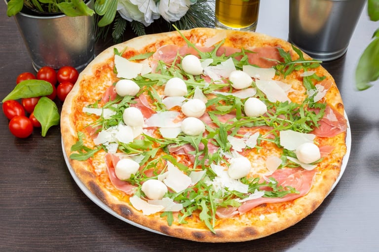 Gourmet pizza with prosciutto, mozzarella, fresh arugula, and herbs on white plate with fresh vegetables and drinks nearby