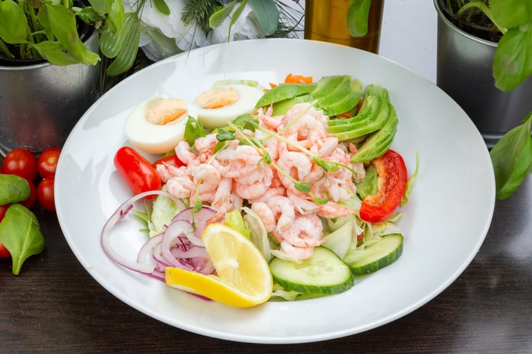 Plate of shrimp salad with fresh vegetables, avocado, boiled eggs, and lemon wedge on white plate