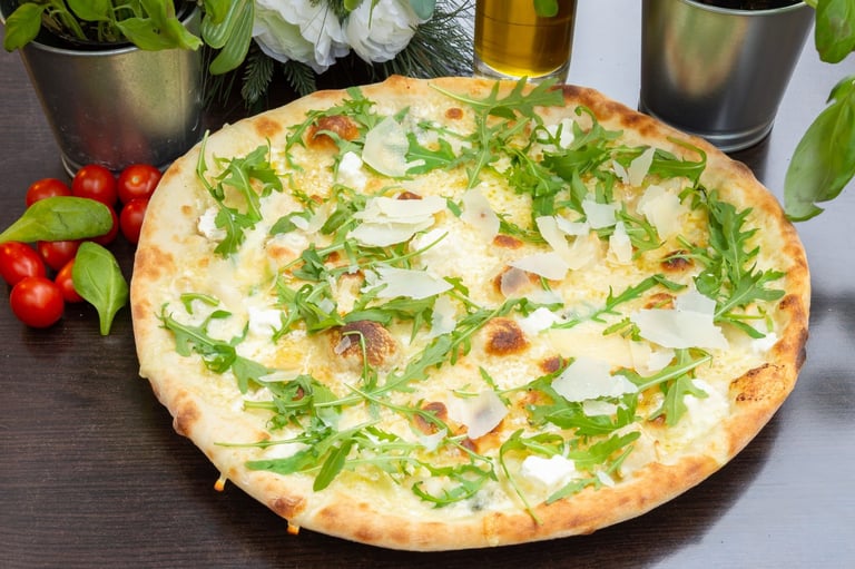 Freshly baked pizza topped with white cheese, arugula, and herbs on dark surface surrounded by fresh vegetables and potted plants